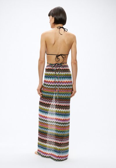 Ženska stoji bosa, obrnjena stran, oblečena v zavezano črno bikini zgornji del in dolgo, barvno krilo z vzorcem cikcak črt.