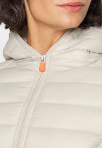 Hellbeige Daunenjacke mit Kapuze, die horizontale gesteppten Bereiche und einen silbernen Reißverschluss mit orangefarbenem Zugband aufweist. Glatte Textur.