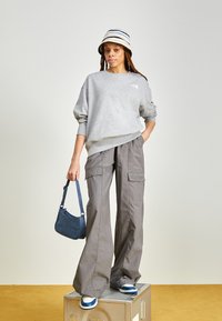 Felpa grigia in pile, pantaloni cargo beige larghi, cappello a secchiello a righe, borsa blu e scarpe da ginnastica blu e bianche su una superficie metallica.