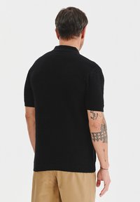 Czarna koszulka polo z krótkim rękawem, materiał teksturowany, ściągacz przy kołnierzyku i dołem, noszona z jasnymi spodniami, widoczne tatuaże na ramieniu.