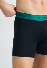 Pantaloni boxer neri con un elastico verde in vita che presenta il logo "Calvin Klein". Texture liscia, forma aderente e lunghezza fino alla metà della coscia.