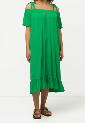 Groene off-shoulder midi-jurk met ruches aan de zoom en strikbandjes, gedragen met bruine open-toe sandalen met gespen, tegen een effen witte achtergrond.