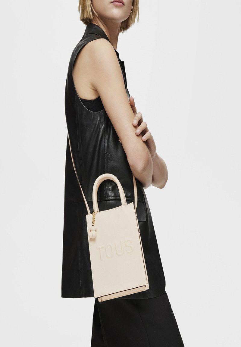 Bolso de mano de cuero color rosa claro con el logo embossed de "TOUS". Cuenta con asas cortas, una correa desmontable y un detalle decorativo de pom-pom.