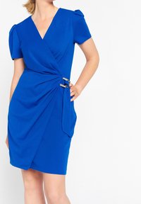 Blå omslagklänning i ett mjukt tyg. Har korta ärmar, en djup V-ringning, sidofyrkningar och en guldbuckle detalj i midjan.