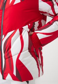 Veste en duvet rouge et blanche avec des motifs ondulés audacieux, fermeture éclair et poignets élastiqués pour un ajustement parfait. Texture douce et matelassée.