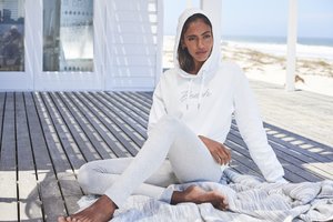 Frau in weißem Hoodie und hellgrauen Leggings sitzt barfuß auf einer Decke auf dem Holzdeck eines Strandhauses, sandiger Uferbereich sichtbar.