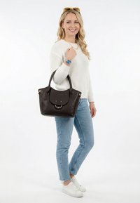 Noor naine valges kampsunis ja teksades, hoides tumepruuni nahast käekotti, kandes valgeid tossusid ja peas kollaseid päikeseprille.