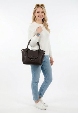 Mujer joven con suéter blanco y jeans, sosteniendo un bolso de cuero marrón oscuro, lleva zapatillas blancas y gafas de sol amarillas en la cabeza.