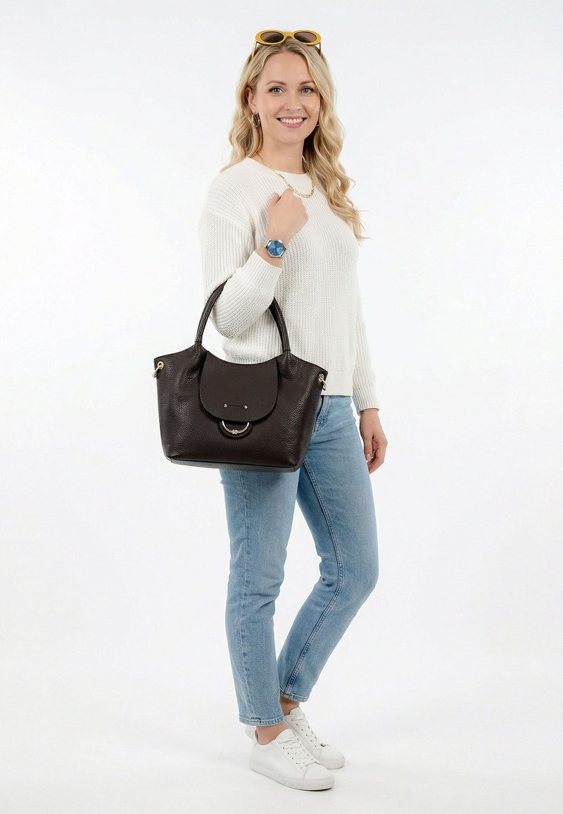 Noor naine valges kampsunis ja teksades, hoides tumepruuni nahast käekotti, kandes valgeid tossusid ja peas kollaseid päikeseprille.