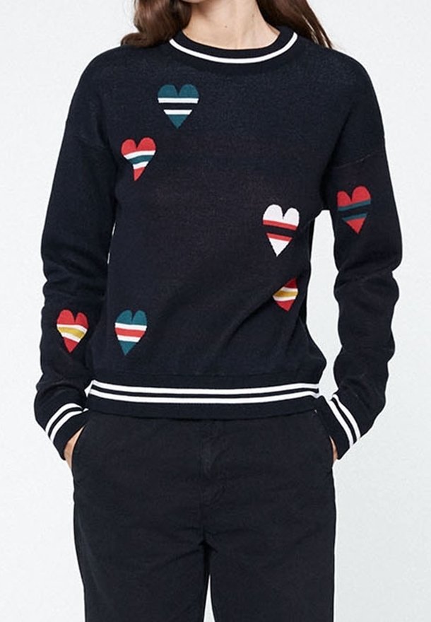 Personne portant un pull sombre avec des motifs de cœurs rayés colorés, les mains dans les poches, et un pantalon noir assorti, vue du cou aux hanches.