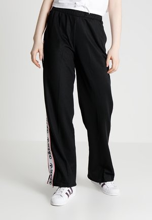 Osoba ubrana w czarne spodnie dresowe Adidas z bocznymi paskami z logo oraz białe sneakersy z ciemnymi paskami, stojąca na białej podłodze.