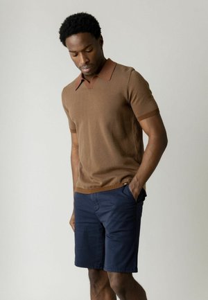 Mężczyzna w brązowej koszulce polo z krótkim rękawem i granatowych krótkich spodenkach, stojący z jedną ręką w kieszeni, patrzący w dół na jednolitym tle.