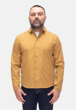 Uomo con capelli corti e barba, indossa una maglia a maniche lunghe color senape e jeans blu scuro, in piedi davanti a uno sfondo neutro.