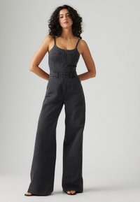 Črni denim kombinezoni z oprijetim bodijem, širokimi nogavicami in zapiranjem na zadrgo spredaj. Ima tanke naramnice in minimalističen dizajn.