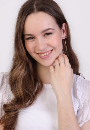 Silberne Creolen und geschichtete silberne Ringe. Das Model hat lange braune Haare, trägt ein geripptes weißes Oberteil mit Rüschenärmeln und lächelt.