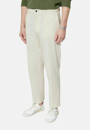 Mężczyzna ubrany w kremowe spodnie o prostych nogawkach, białe trampki, zieloną koszulę z długim rękawem oraz srebrne bransoletki na prawej ręce, stojący na jednolitym tle.