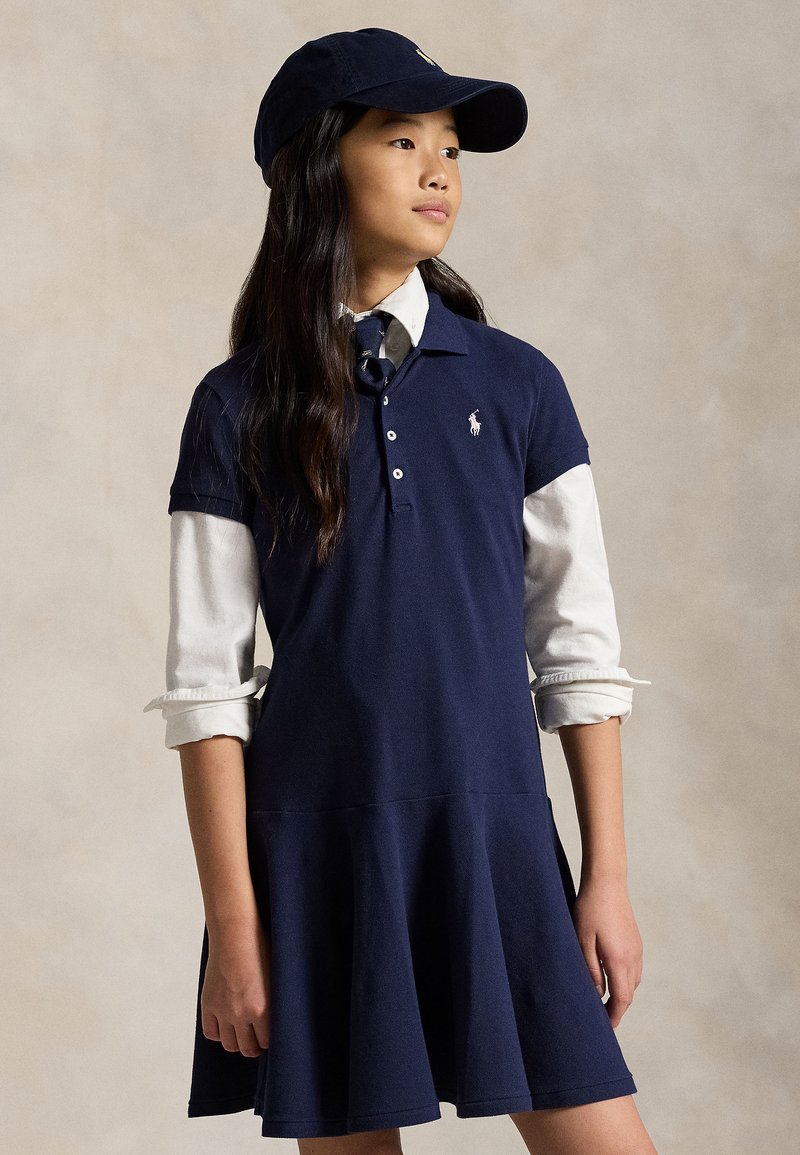 Giovane ragazza che indossa un berretto blu navy e un vestito polo sopra una camicia bianca a maniche lunghe, in piedi davanti a uno sfondo beige uniforme.