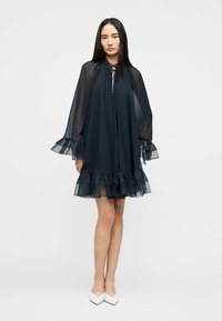 Frau trägt ein locker sitzendes schwarzes, transparentes Kleid mit gerafften Manschetten und Saum, steht mit entspannten Armen, kombiniert mit weißen spitzen High Heels.