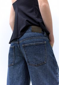 Tummansiniset farkut, joissa on teksturoitu pinta, korkea vyötärö ja isot takataskut. Vyötäröllä on ruskea nahkalappu.