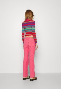 Haut à manches longues rayé rouge, vert et bleu, associé à un pantalon évasé en velours côtelé rose, avec une étiquette à la taille et des chaussures décontractées.