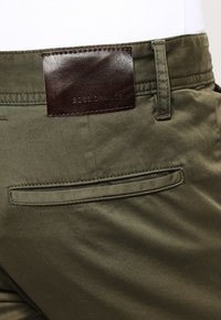 Olivgröna byxor med en läderetikett med präglad text, en bakficka och slät tygtextur.