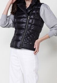 Femme portant une veste matelassée noire sur une chemise à manches longues gris clair et un pantalon blanc, les mains reposant près de son visage et de sa hanche.