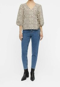 Blouse à manches ballon en crème avec un motif en grille texturé, présentant un décolleté en V profond. Associée à un jean bleu taille haute et des bottines noires.