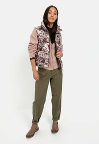 Blomsterpræget ydervest over en let sweater, parret med olivengrønne bukser og brune ankelstøvler. Glatte stoffer og tætsiddende design.