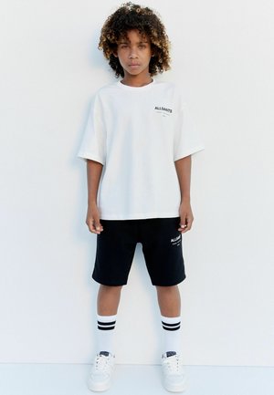 Weißes Oversize-T-Shirt mit "ALLSAINTS"-Logo, kombiniert mit schwarzen Shorts, weißen Sneakern und weißen Socken mit schwarzen Streifen. Lockige Haarstruktur.