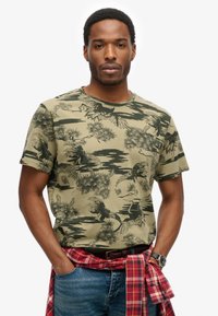 Mand i grøn mønstret t-shirt med fugle- og trædesign, blå jeans, rødt ternet skjorte bundet om livet og armbåndsur.