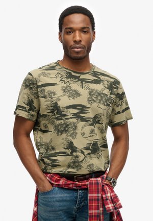 Man in een groene T-shirt met een patroon van vogels en bomen, blauwe jeans, een rode geruite shirt om zijn taille geknoopt, en een horloge.