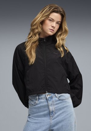 Zwarte cropped windbreakerjas met een hoge kraag, ritssluiting en gestructureerde stof, gecombineerd met lichtblauwe spijkerbroek.