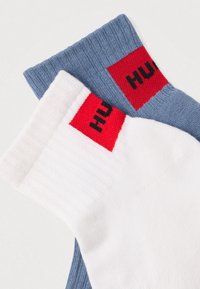 Două perechi de șosete: una albă cu o bandă roșie cu text negru, cealaltă albastru deschis cu o bandă roșie. Ambele au textură reiată.