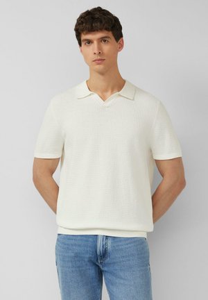 Mężczyzna z krótkimi kręconymi włosami, ubrany w białą teksturowaną koszulkę polo z krótkim rękawem i niebieskie dżinsy, stoi na tle gładkiej ściany.