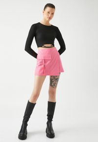 Mustapintainen pitkähihainen crop-top, jossa on ribattu pinta, yhdistettynä vaaleanpunaiseen cargo-skorttiin, jossa on suuri sivutasku. Mustat polvipituiset saappaat.