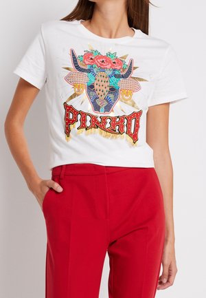 Kvinde iført en hvid t-shirt med et farverigt tyrehovede og "Pincho" -tekstdesign, kombineret med højtaljede røde bukser.