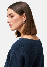 Granatowy sweter dzianinowy o ribbowej fakturze z rozproszonymi akcentami błyszczącymi. Okrągły dekolt, luźny krój oraz opadające ramiona.