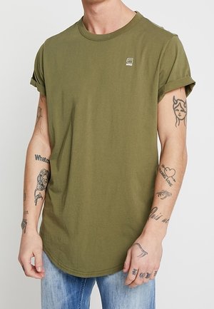 Homme portant un t-shirt vert olive à manches retroussées et un jean bleu clair, exhibant des tatouages sur les deux avant-bras et les mains sur un fond uni.