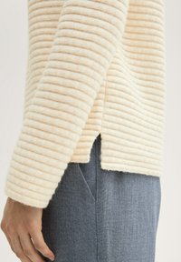 Pull en crème rayé en tissu doux, avec une texture côtelée, un ourlet fendu et des manches ajustées, associé à un pantalon en jean foncé.