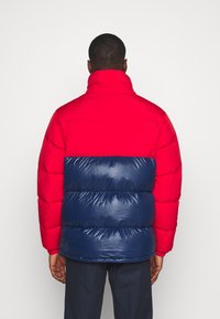 Vadderad jacka med stark röd överdel och marinblå midja. Har hög krage och glänsande yta, med synlig sömnad.