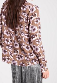 Top floreale marrone con motivi di fiori viola e crema, maniche lunghe, vestibilità comoda, abbinato a pantaloni plissettati metallici argento.