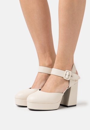 Beige Mary Jane-platformpumps met dikke blokhakken en enkelbandjes met vierkante gespen aan iemands voeten.