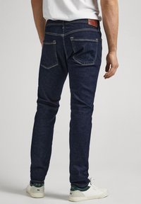 Hombre vistiendo jeans slim-fit azul oscuro, zapatillas blancas y una camiseta blanca, de pie mirando hacia atrás contra un fondo liso.