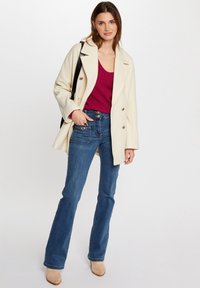Cappotto crema con design doppiopetto, maglione a maglia berry e jeans blu svasati; presenta dettagli unici nelle tasche e nei bottoni. Stivali beige con tacco.