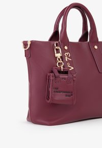Bolso tote de cuero burdeos con asas dobles, herrajes dorados y un pequeño bolsillo adjunto que muestra "SOY UNA BOLSA RESPONSABLE."