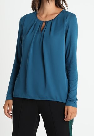 Femme portant un chemisier à manches longues bleu sarcelle avec une petite ouverture devant et un pantalon noir, debout sur un fond blanc.