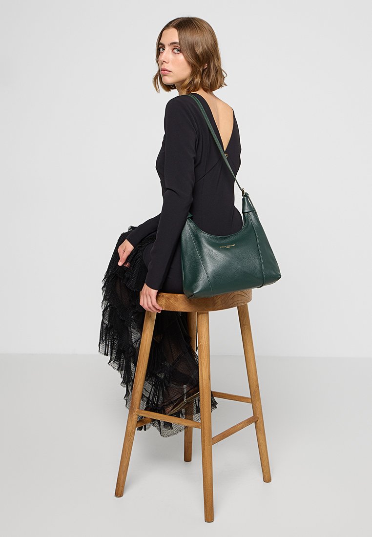 Mujer con cabello corto castaño, vestida con un vestido negro de falda de encaje, sentada en un taburete de madera, llevando un bolso de hombro de cuero verde oscuro.