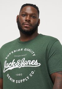 Grüne Baumwoll-T-Shirt mit einem weißen Logo und dem Schriftzug "Jack & Jones" sowie "Superior Quality" auf der Vorderseite. Rundhals und kurze Ärmel.