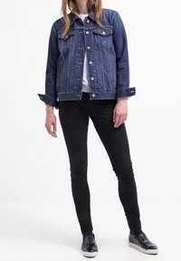 Chaqueta de mezclilla azul oscura con bolsillos frontales y botones plateados, combinada con jeans skinny negros y zapatos negros sin cordones.