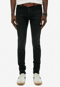 Vaqueros de mezclilla negra con corte slim, cinco bolsillos y un cinturón de cuero marrón. Combinados con zapatillas blancas con acentos negros.